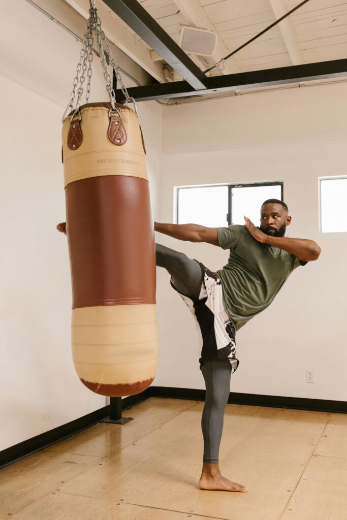 pexels photo 8612529 8612529 A man practicing martial arts with a high kick on a punching bag indoors.